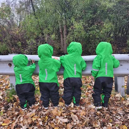 Four children in green rain suits looking over a guardrail into the woods, backs to camera