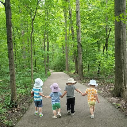 Four children holding hands walking down a woodland path in sun hats