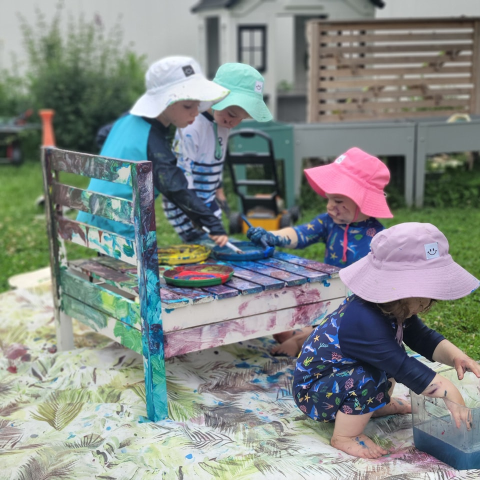 Children painting at an outdoor bench wearing sun hats