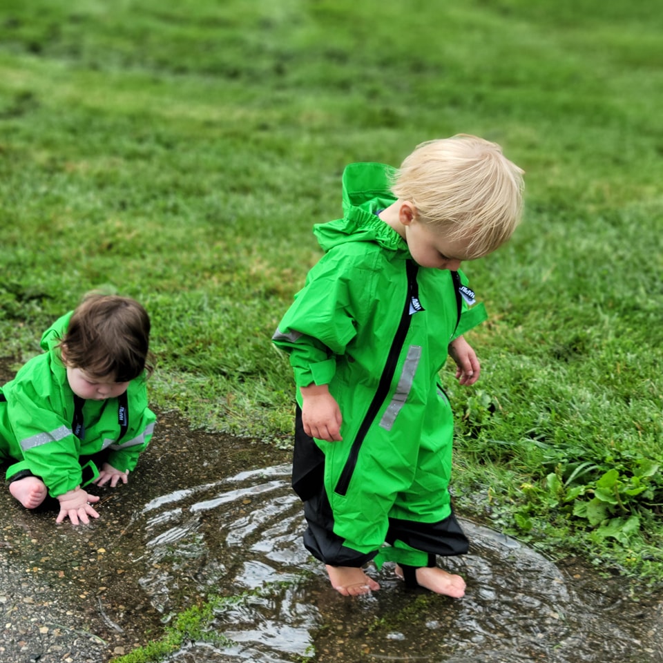 Children in green rain suits splashing barefoot in a puddle
