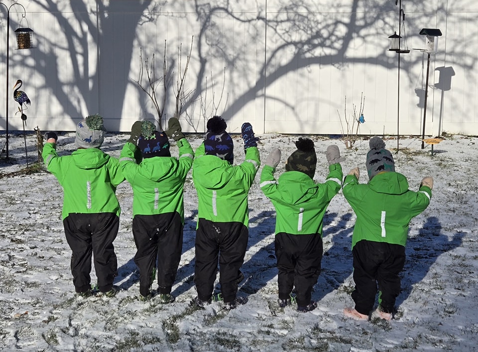 Six children in green jackets standing in the snow with backs to camera, arms raised